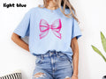 Light Blue: Light blue t-shirt with a bright pink ribbon bow graphic worn by a model perfect for a coquette aesthetic gift