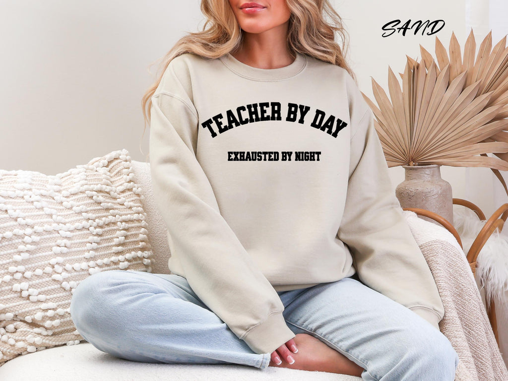 Sand: Woman sitting wearing a sand teacher by day exhausted by night sweatshirt which makes a perfect teacher gift
