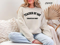 Sand: Woman sitting wearing a sand teacher by day exhausted by night sweatshirt which makes a perfect teacher gift