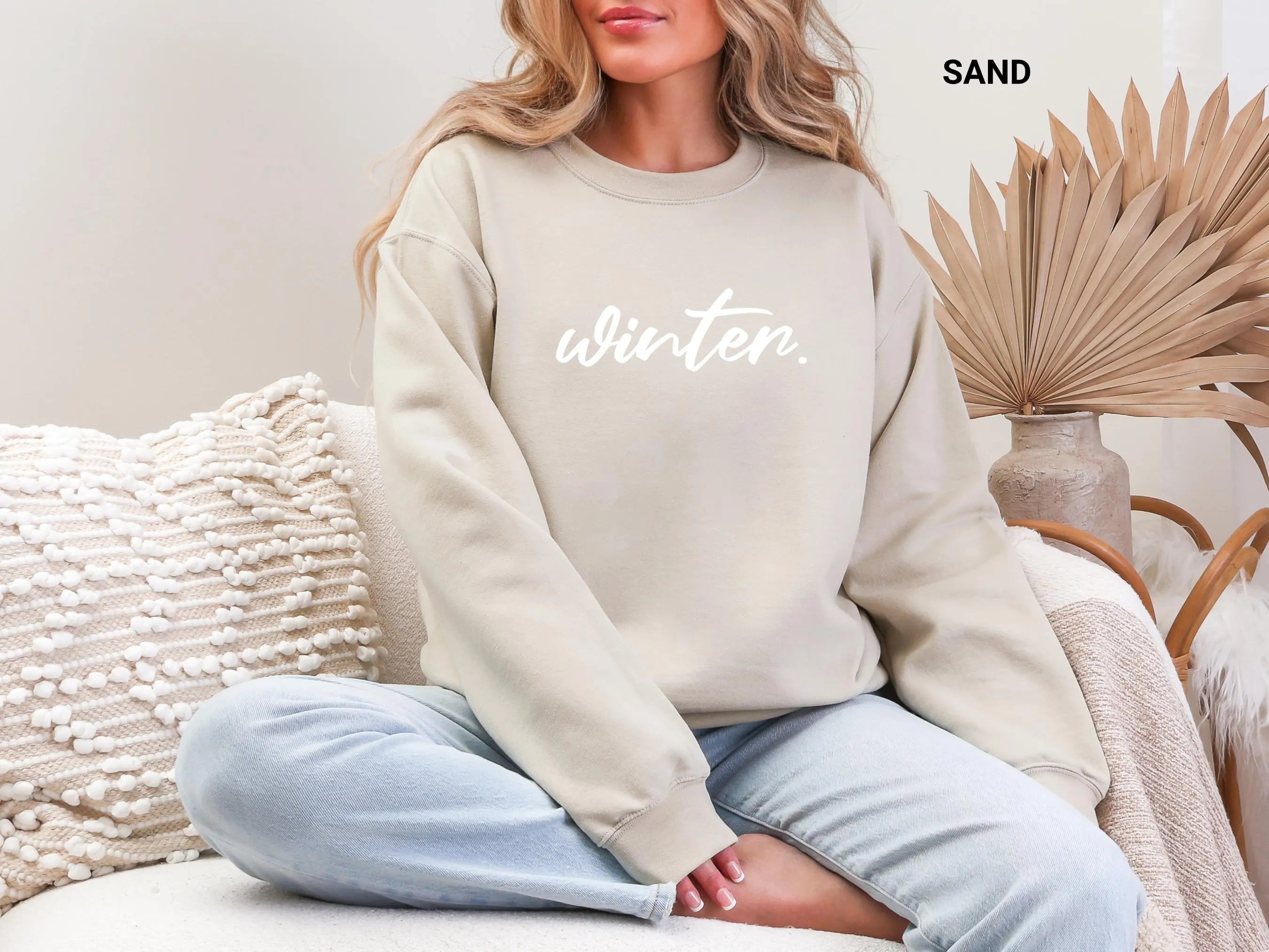 Sand: Woman wearing a cozy sand sweatshirt with the word winter in white cursive lettering sitting on a textured sofa