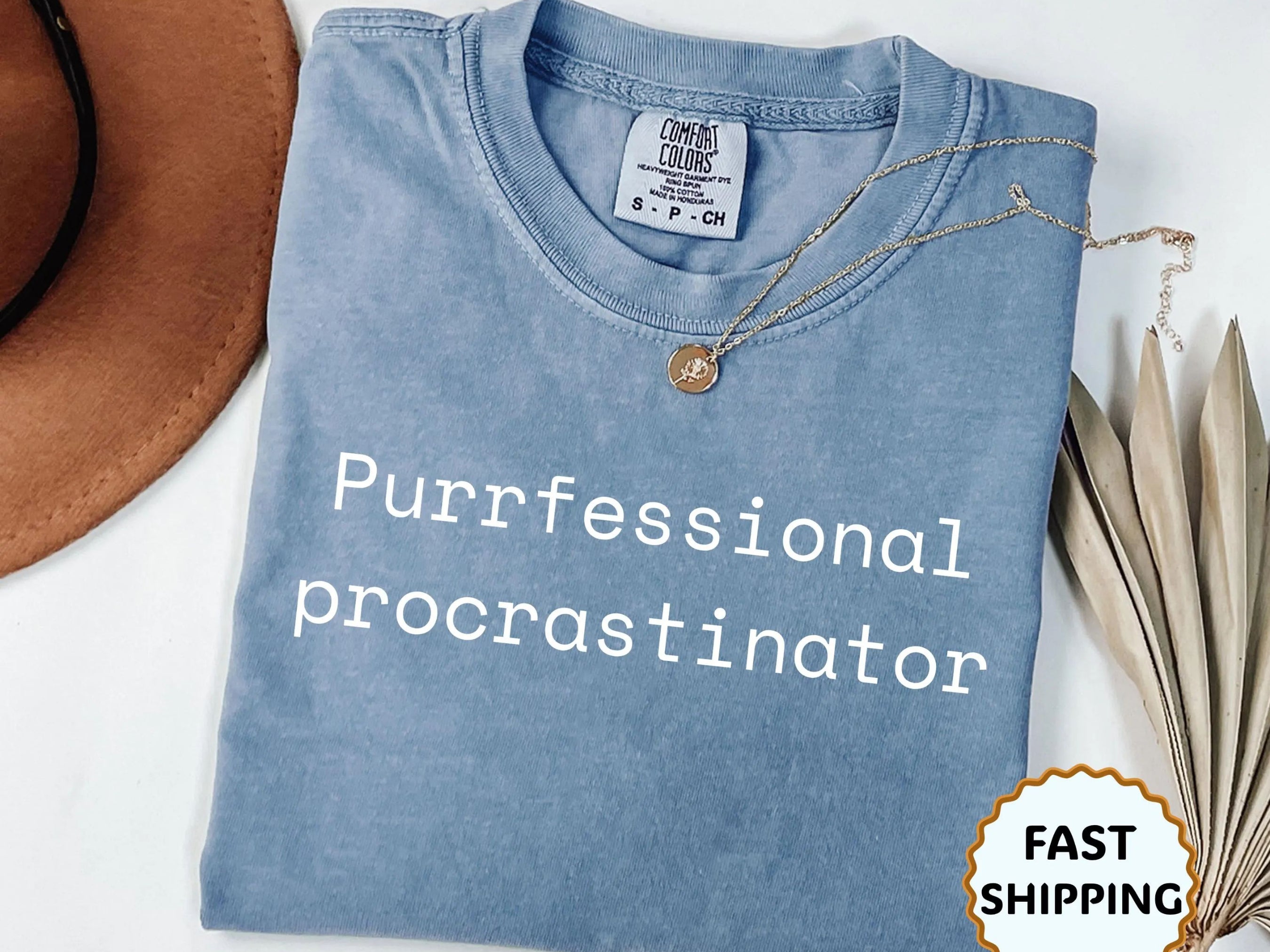 Folded blue t-shirt with 'Purfessional procrastinator' text, necklace, and hat on a white background