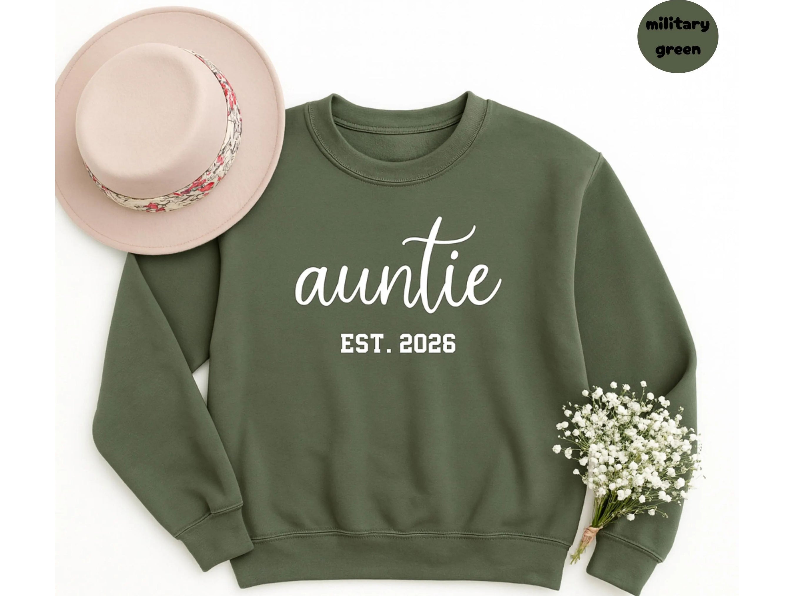 Military green sweatshirt with 'auntie est. 2026' text, pink hat, and white flowers on a light background