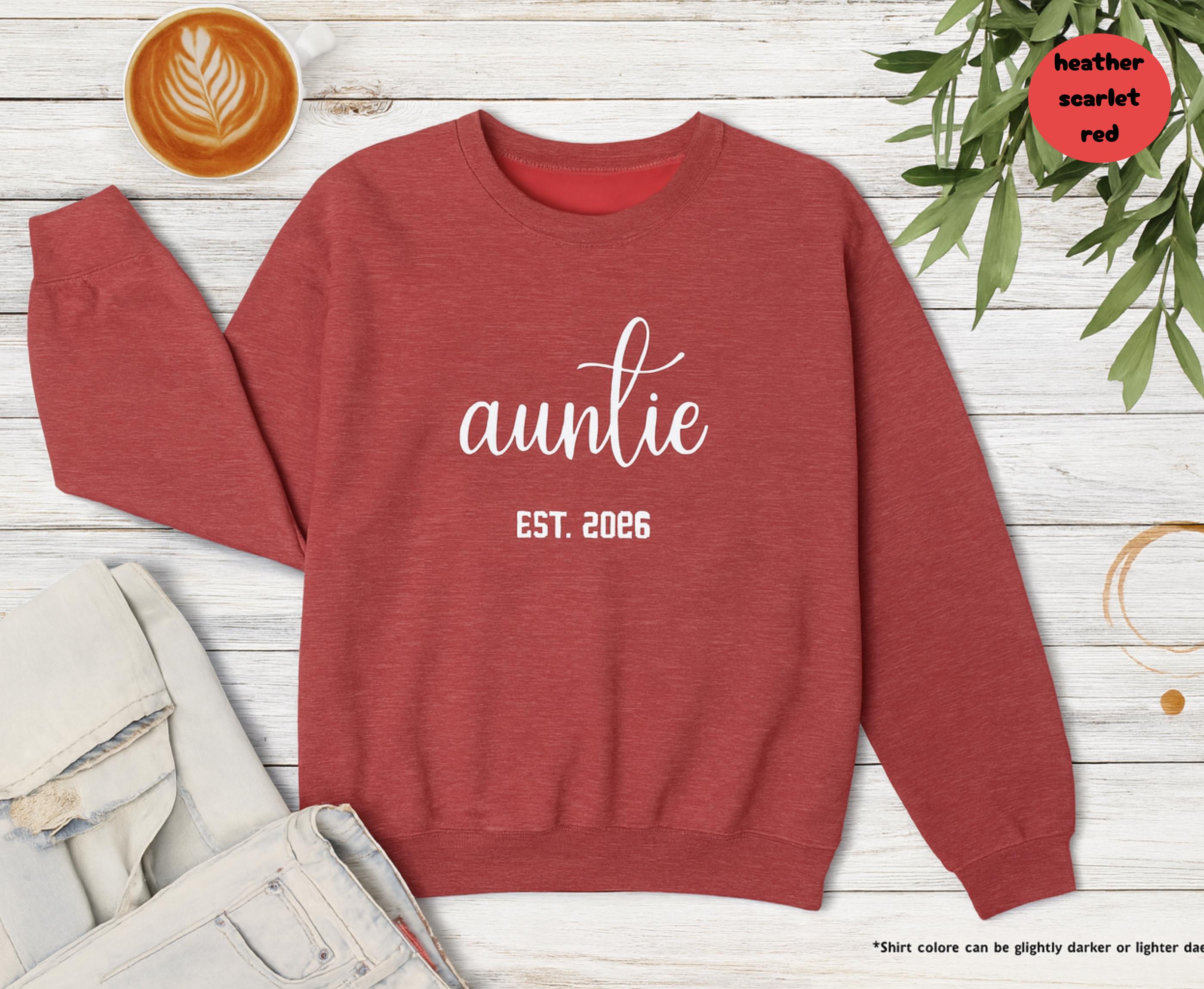 Heather scarlet red Auntie Est 2026 sweatshirt folded on a wooden table next to coffee and leaves