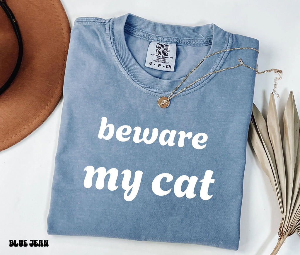 Folded blue shirt with 'beware my cat' text on a white surface with a hat and plant.