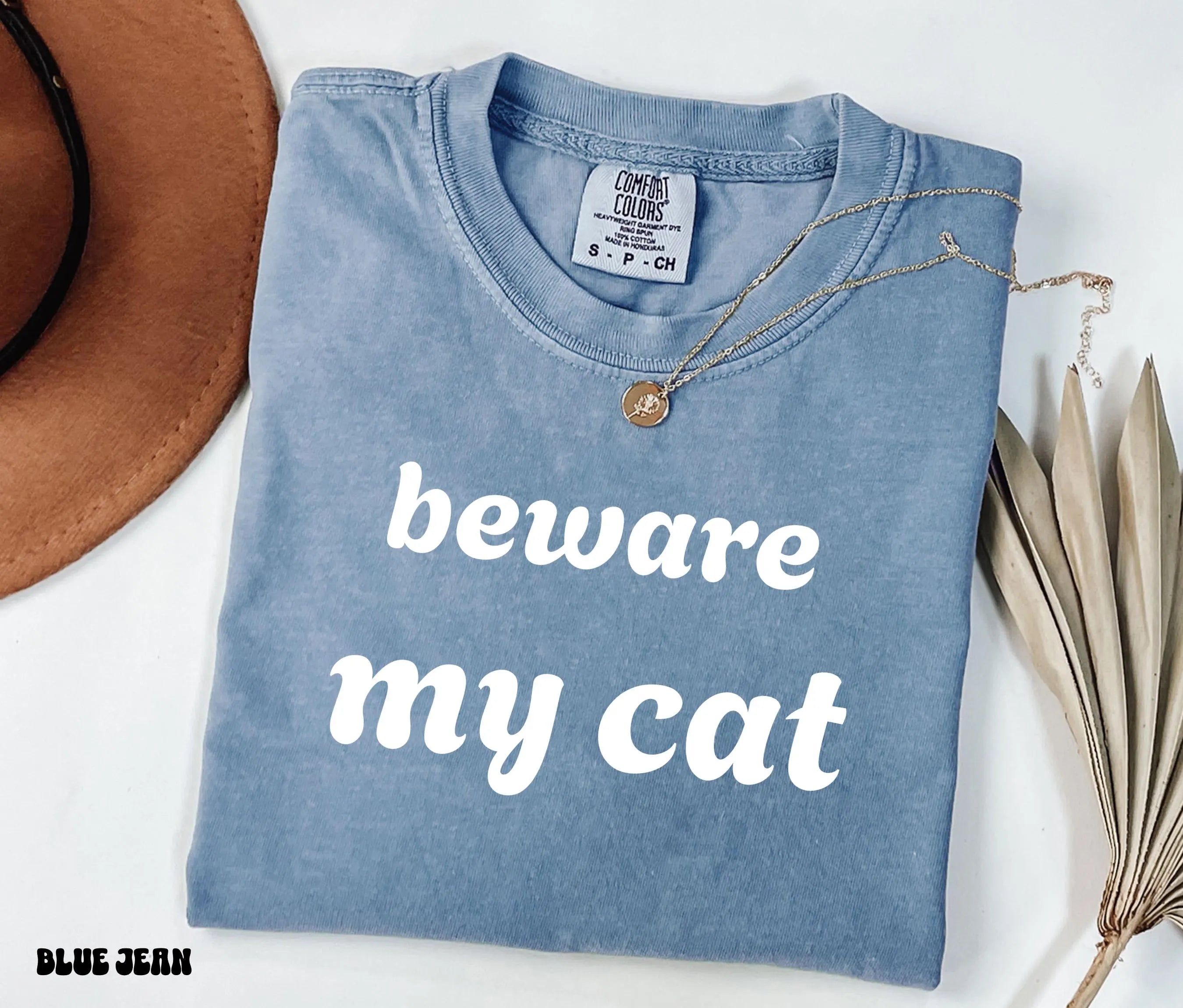 Folded blue shirt with 'beware my cat' text on a white surface with a hat and plant.