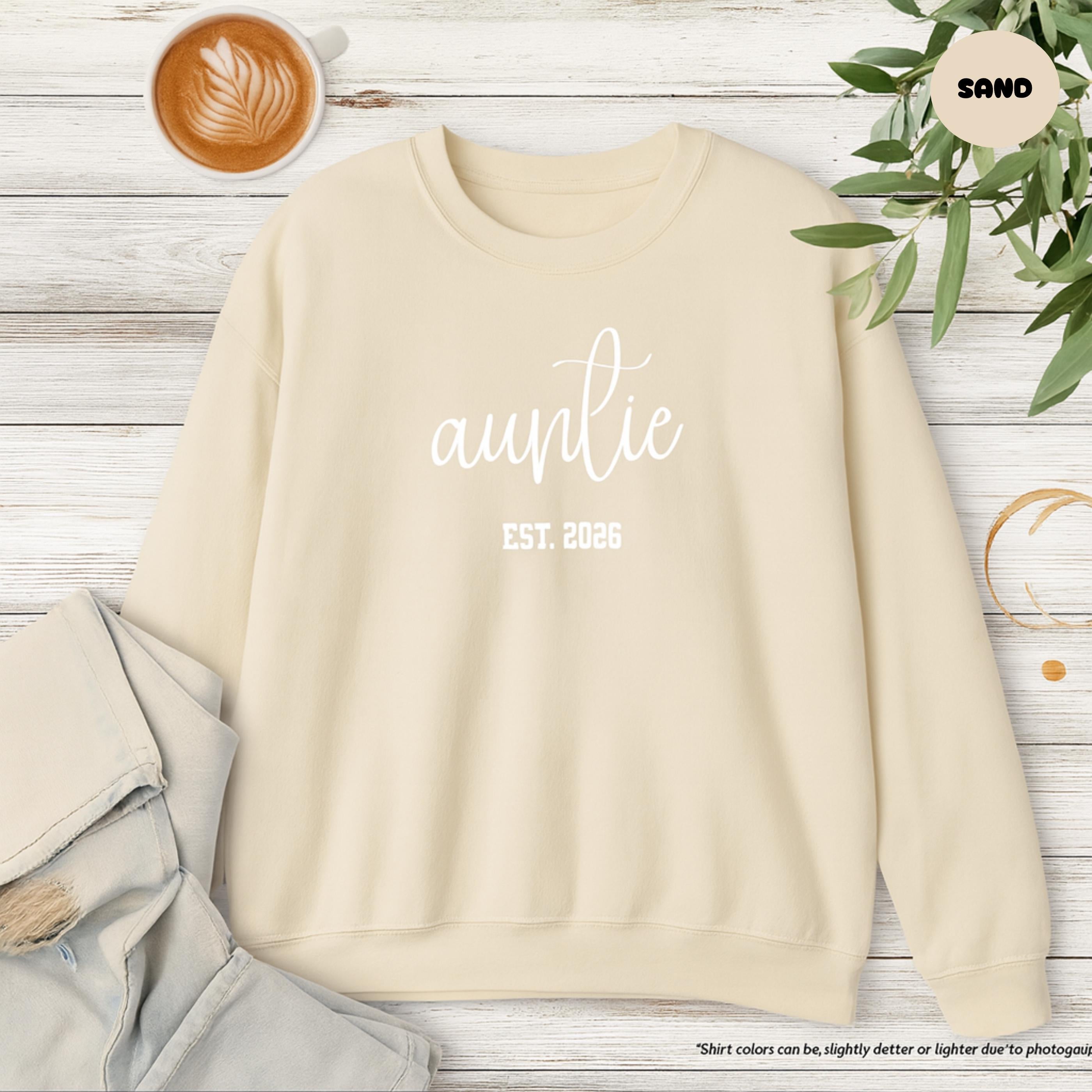 Beige sweatshirt with 'auntie est. 2026' text on a wooden surface with a coffee cup and plant.