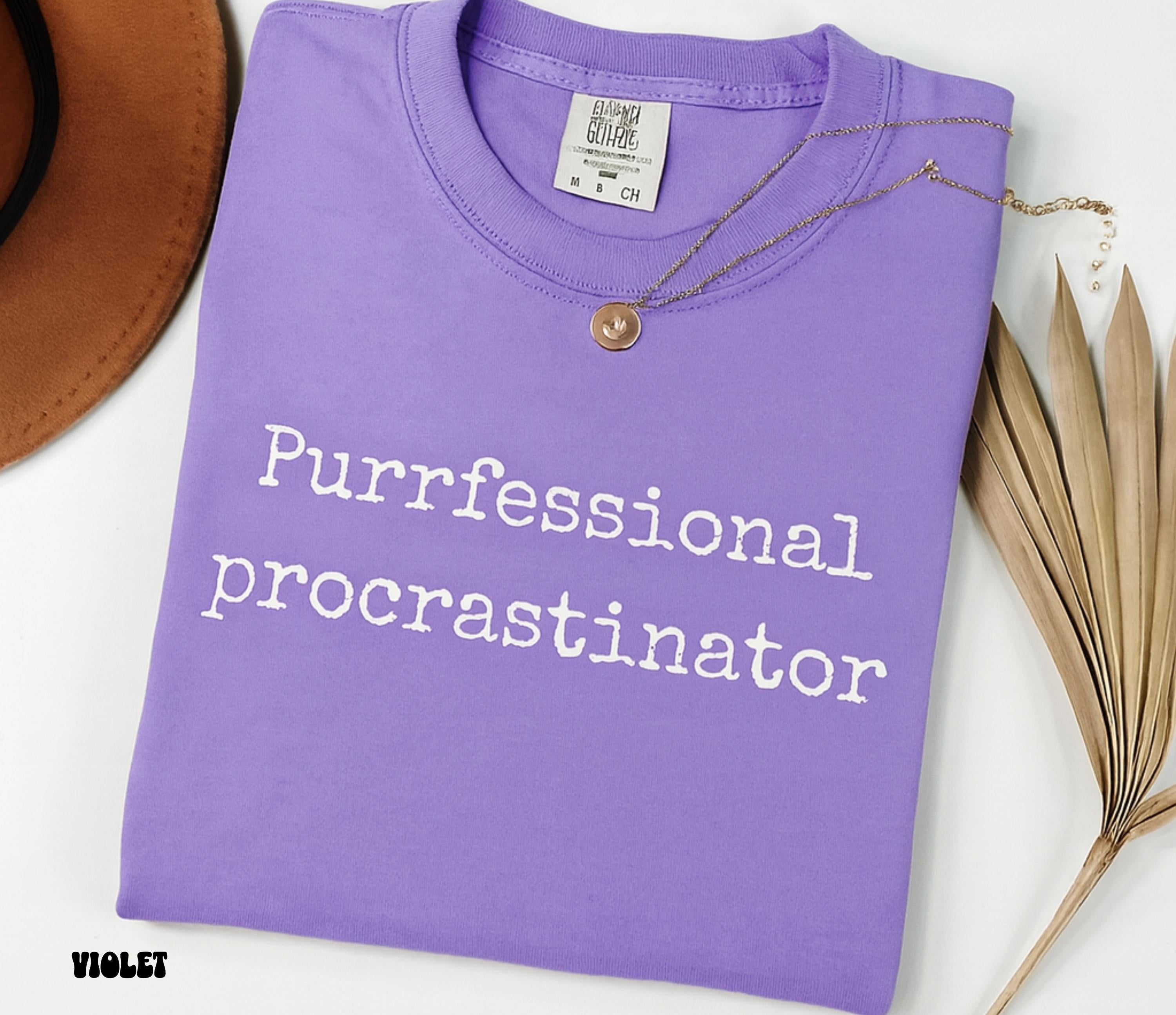 Folded purple t-shirt with 'Purrfessional procrastinator' text on a white surface.