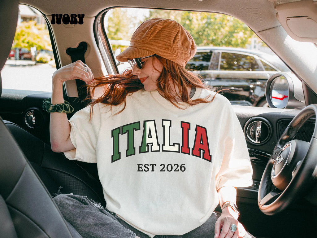 Woman wearing ivory Italia Est 2026 collegiate t shirt with green white and red text sitting in car